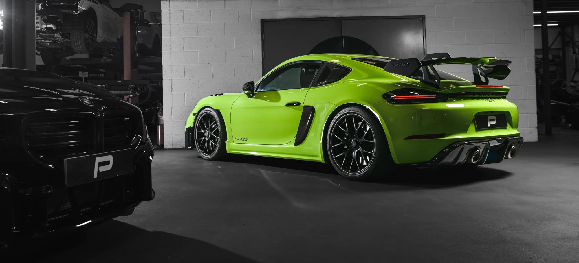 Porsche 718 GT4 RS with Akrapovic exhaust installed, parked in PERON's Performance Workshop in front of the in-house dyno, with a G87 BMW M2 in the foreground.