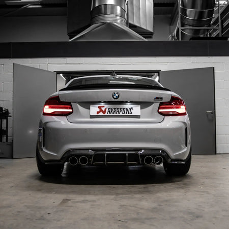 Rear view of the F87N BMW M2 in PERON's Performance Workshop, highlighting the installed Akrapovic exhaust system with carbon tailpipes.