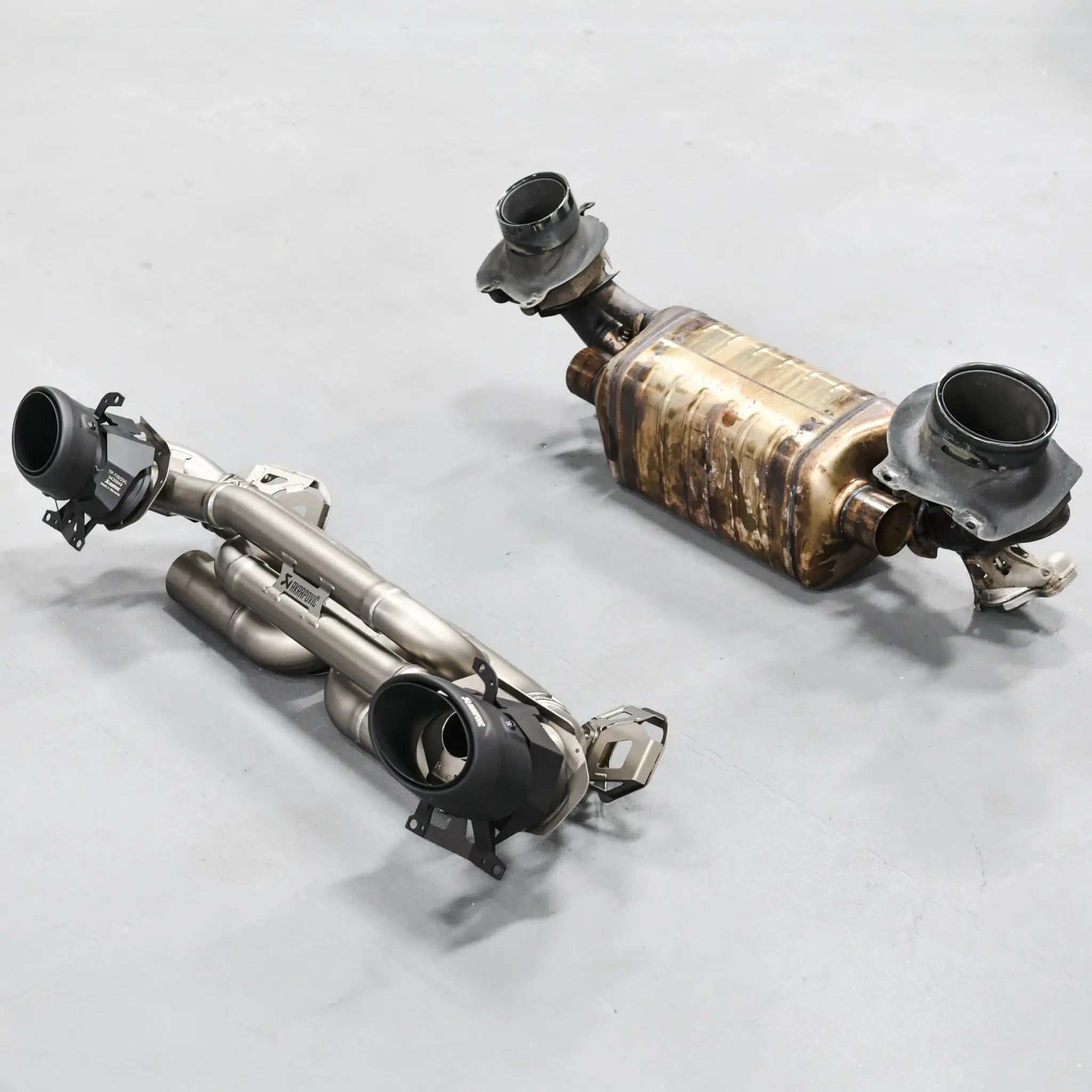 Side-by-side comparison of the Akrapovič Slip-On Race Line (Titanium) performance exhaust system and the stock Porsche 911 Turbo S 992.1 exhaust system, showcasing weight and design differences.